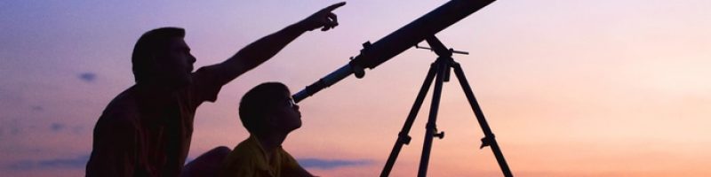 A young boy looking thru a telescope at sunset with his father.